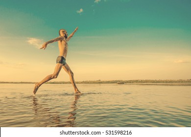 Adult male with a mohawk on his head and black shorts running on water against the backdrop of blue sky with clouds in the sunlight