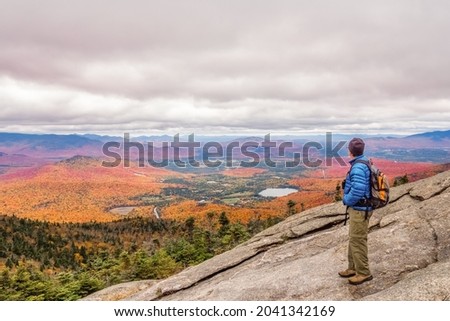 Adult male on top of mountain in Adirondacks looks at fall foliage under moody sky on cold day.