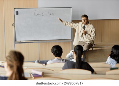 Adult male professor of law lecturing students while sitting on desk pointing at writing on whiteboard in university classroom, copy space