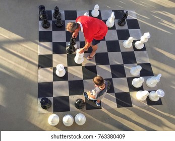 Adult Male Takes Bishop With Rook During Game Of Chess Against A Boy On Giant Outdoor Chessboard, Viewed From Above. Outdoor Chess Game Provides Entertainment On Deck Of Cruise Ship