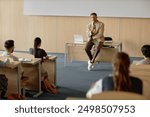 Adult male teacher with hand held microphone lecturing university students while leaning on desk in lecture hall, copy space