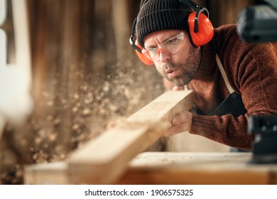 Adult male woodworker in protective goggles and headphones blowing sawdust from wooden detail while working in carpentry workshop - Powered by Shutterstock