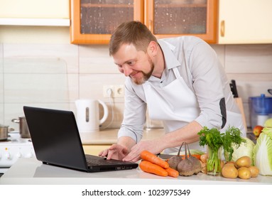 Adult Man Of 40 Years Old Prepares A Vegetable Dish In The Kitchen On A Prescription From The Internet. Watch In The Laptop And Cook Food. Vegetarian Menu In The Diet - Taking Care Of Your Health.