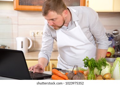 Adult Man Of 40 Years Old Prepares A Vegetable Dish In The Kitchen On A Prescription From The Internet. Watch In The Laptop And Cook Food. Vegetarian Menu In The Diet - Taking Care Of Your Health.