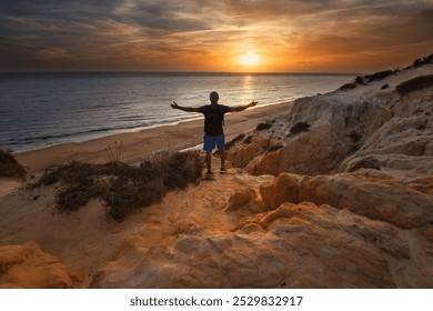 An adult man, adventurer, traveler with a backpack, standing on the edge of a cliff, facing the sea. Opening his arms, breathing deeply, contemplating the sunset. Meditation, well-being, retired. - Powered by Shutterstock