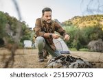 Adult man with a backpack and hiking attire smiles while prepare sticks for a campfire at a scenic outdoor campsite with a tent and chair in the background