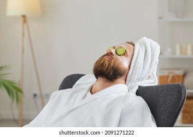 Adult man in bathrobe and towel turban enjoying spa day at home and relaxing in comfortable chair or armchair with facial beauty mask on face and fresh cucumber slices on eyes. Skin care concept - Shutterstock ID 2091271222