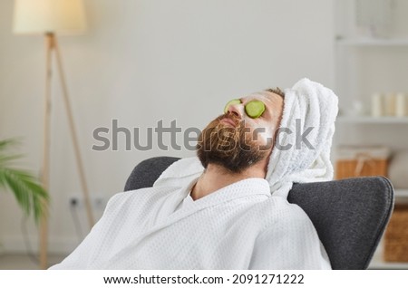 Adult man in bathrobe and towel turban enjoying spa day at home and relaxing in comfortable chair or armchair with facial beauty mask on face and fresh cucumber slices on eyes. Skin care concept