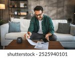 Adult man in casual attire sit on a sofa, holding a bill and a smart phone, with other documents on the table and a coffee mug, focusing on managing his finances