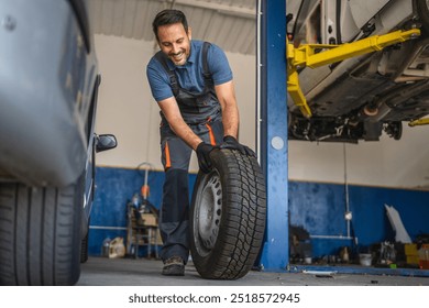 Adult man cheerful car mechanic in a blue shirt and gloves handles a tire confidently at an auto repair garage, showcasing professionalism and automotive expertise - Powered by Shutterstock