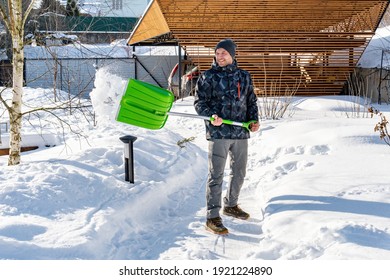 An adult man cleans the paths in the garden from snow in a good mood. Shoveling snow from paths with a shovel on a sunny day. Garden care in winter. Snow falls off the shovel when cleaning snow.