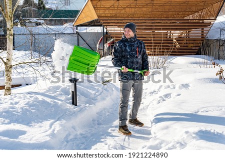 An adult man cleans the paths in the garden from snow in a good mood. Shoveling snow from paths with a shovel on a sunny day. Garden care in winter. Snow falls off the shovel when cleaning snow.