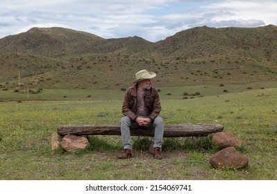 Adult Man In Cowboy Hat Sitting On Wooden Bench In Field