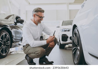 Adult man customer buyer client wear shirt take photo on mobile cell phone wheel rim tire choose auto want buy new automobile in car showroom vehicle salon dealership store motor show. Sales concept - Powered by Shutterstock