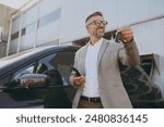 Adult man customer male buyer client wearing white shirt classic grey suit standing near black electric car while charging looking aside use mobile cell phone hold give keys outside. Business concept