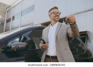 Adult man customer male buyer client wearing white shirt classic grey suit standing near black electric car while charging looking aside use mobile cell phone hold give keys outside. Business concept - Powered by Shutterstock