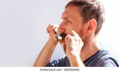 Adult Man Cutting His Own Beard And Mustache With Scissors And Comb. Caucasian Red Bearded Male Trimming Hair On Face At Home. Do It Yourself Self-made Haircut.Selfcare DIY During Quarantine Isolation