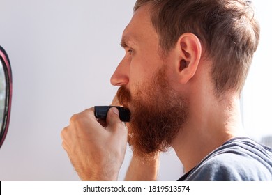 Adult Man Cutting His Own Beard And Mustache With Scissors And Comb. Caucasian Red Bearded Male Trimming Hair On Face At Home. Do It Yourself Self-made Haircut.Selfcare DIY During Quarantine Isolation