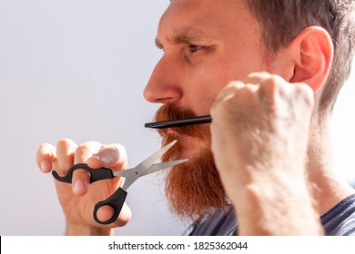 Adult Man Cutting His Own Beard And Mustache With Scissors And Comb. Caucasian Red Bearded Male Trimming Hair On Face At Home. Do It Yourself Self-made Haircut.Selfcare DIY During Quarantine Isolation