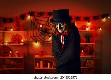 Adult man dressed up as evil vampire or sinister maniac for macabre Halloween party. Guy with long ginger hair in top hat and cloak looking at camera standing in dark interior with orange red lights