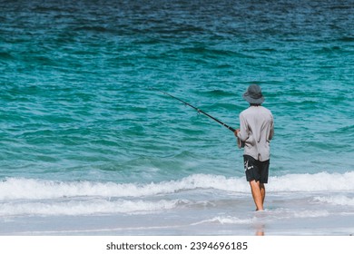 adult man fishing with his fishing rod and his hat on his head, on the seashore on the white sand beach and turquoise waters on a cloudy day - Powered by Shutterstock