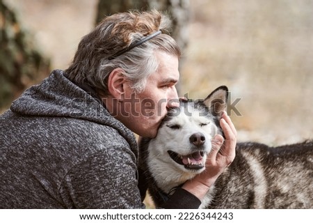 Adult man in gray sweatshirt hugs and kisses Siberian Husky dog, true love of human and pet, funny meet of black white Husky dog and owner. Man kisses Husky dog outdoor forest background