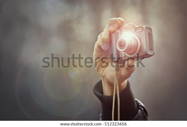 Adult man hand holding a compact digital\
point-and-shoot photo camera over out of focus outdoor background.\
Taking outdoor pictures with a digital compact camera. Lens flare\
and copy space.