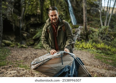 Adult man hiker in a green jacket sets up a camping tent in a scenic forest area with a waterfall in the background, embodying tranquility and adventure spirit. - Powered by Shutterstock