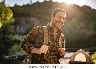 adult man hiker wearing a large backpack on a camping trip, standing near a tent and a folding chair by a river, surrounded by lush forest and hills during sunrise - Powered by Shutterstock