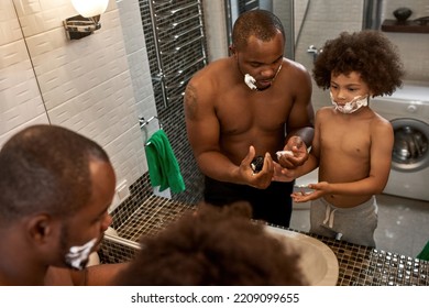 Adult Man Holding Foam And Teaching Little Son Shaving At Home Bathroom. Domestic Family Lifestyle. Black Man And Child Spend Time Together. Fatherhood And Parenting. Morning. Hygiene And Face Care