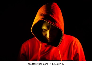 An adult man looks out from under the hood with a grin like a psycho or a maniac in an orange hooded sweatshirt highlighted in red and yellow on the sides on a black isolated background. - Shutterstock ID 1405569449