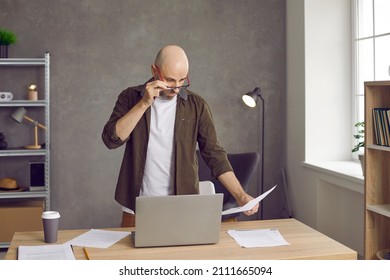 Adult Man Looks In Surprise Through His Glasses At Bills For Payment, Bank Statement Or Debt On Loan. Businessman With Worried Expression Standing At His Desk With Document In Hand In Office.