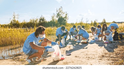 An adult man in mask picking up garbage into trash bag with the group of eco activists in the background. A male volunteer collecting plastic bottles, cardboards, waste. Ecology conservation concept. - Powered by Shutterstock