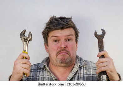 An Adult Man With A Perplexed Face Holds Two Wrenches. A Male With Tangled, Disheveled Hair Faces The Light Gray Wall.  Indoors.  Selective Focus.