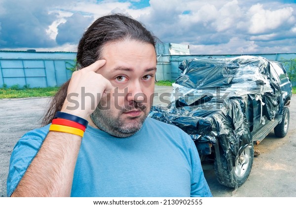 An adult man points his finger at his head, urging\
him to think against the background of a broken car. Concept on the\
theme of foresight and insurance. Bracelet in the colors of the\
German flag.