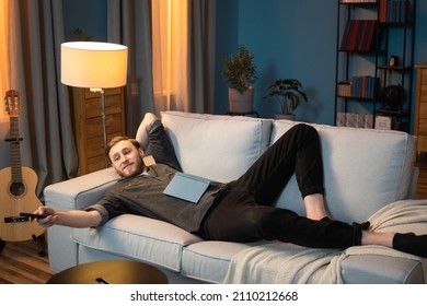 Adult man is resting after work in the evening in living room, lying along the couch, propped up by head, leg bent at the knee, a book lying on stomach, switching channels with the TV remote control