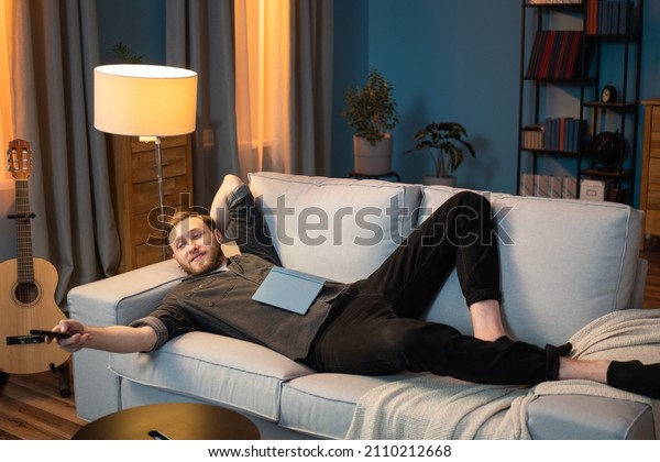 Adult man is resting after work in the evening in\
living room, lying along the couch, propped up by head, leg bent at\
the knee, a book lying on stomach, switching channels with the TV\
remote control