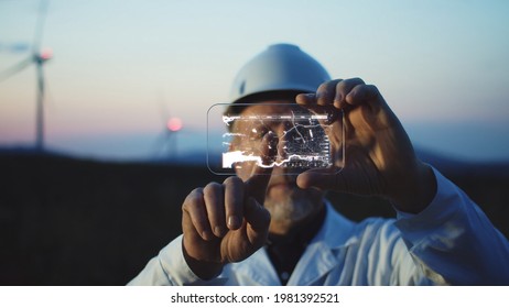 Adult Man Scientist Using High Tech Smart Phone To Diagnose Online The Workflow On Modern Windmill Farm. Beautiful Sunset Sky On Background. The Newest Technology Trend Of Communication.