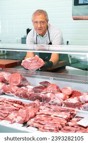 Adult man seller in uniform lays out raw beef in showcase of butcher shop.. - Shutterstock ID 2393282105