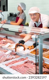 Adult man seller in uniform lays out meat sausages in butcher shop window - Shutterstock ID 2393282197