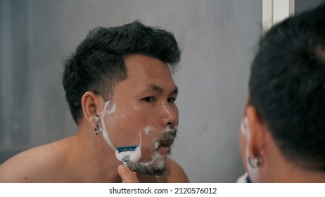 Adult man shaving with foam and manual razer.Mirror reflection young handsome smiling man touching beard, grooming in bathroom. Head shot close up happy asian man guy doing morning beauty routine.