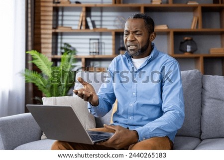 An adult man sits on a sofa with a laptop, showing an expression of frustration and confusion, possibly due to technical issues or bad news.