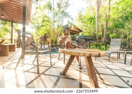 adult man sitting near a wooden table with a coffee in a beach house with a swimming pool in the middle of the tropical forest