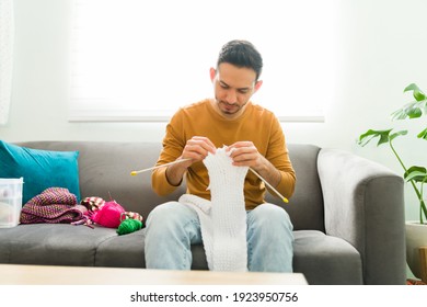 Adult Man Sitting On The Sofa And Knitting A Scarf As Part Of His Mental Health Activities. Hispanic Man Using Needles And Wool Balls To Knit Clothes