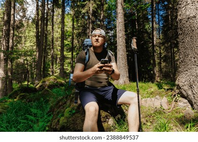 Hombre adulto con mochila para smartphones con bastón de trekking sentado en un bosque verde