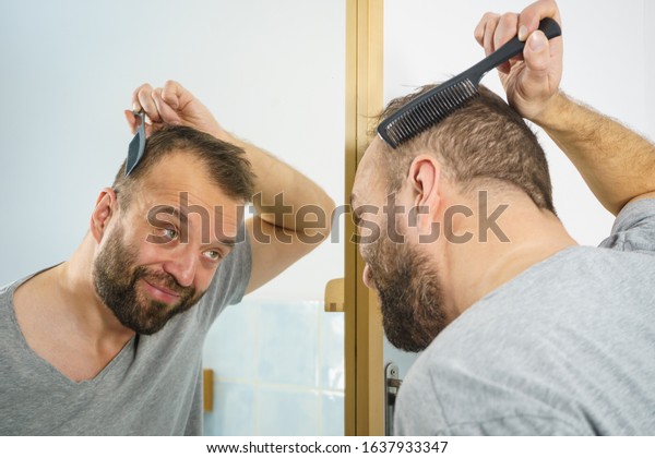 Adult man standing in front of the bathroom\
mirror brushing his short hair using comb. Guy investigating his\
receding hairline