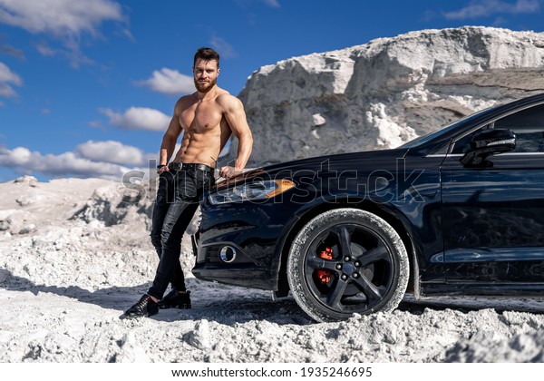 Adult man standing near black modern car and\
holding sunglasses. Attractive man with naked abs. White mountain\
and blue sky on\
background.