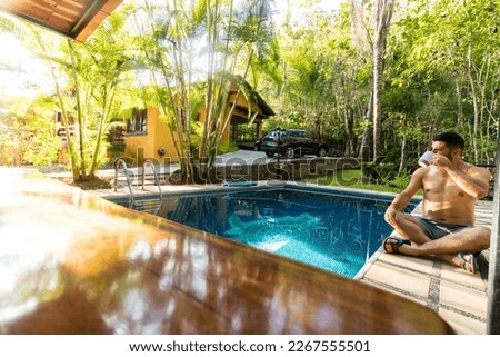 adult man in a swimsuit sitting drinking coffee in a beach house with a swimming pool in the middle of the tropical forest