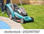 Adult man using a battery powered electric lawnmower to trim grass growing next to the curb of the swimming pool covered with a protective tarp