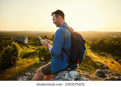 Adult man using phone hiking during sunset sitting on top of the mountain - Powered by Shutterstock